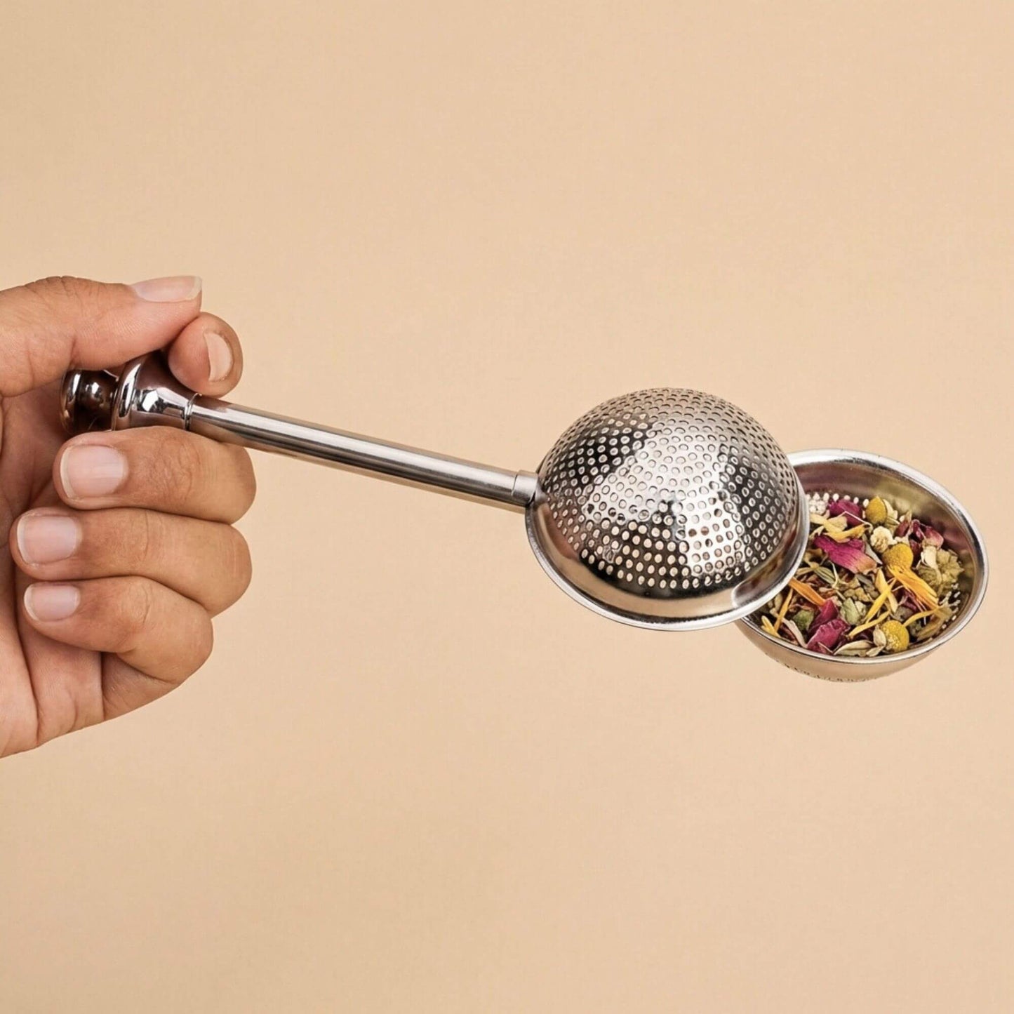 Boule à thé en inox remplie de plantes en vrac, tenue en main, accessoire pour infusion de tisanes et thés sur fond beige.