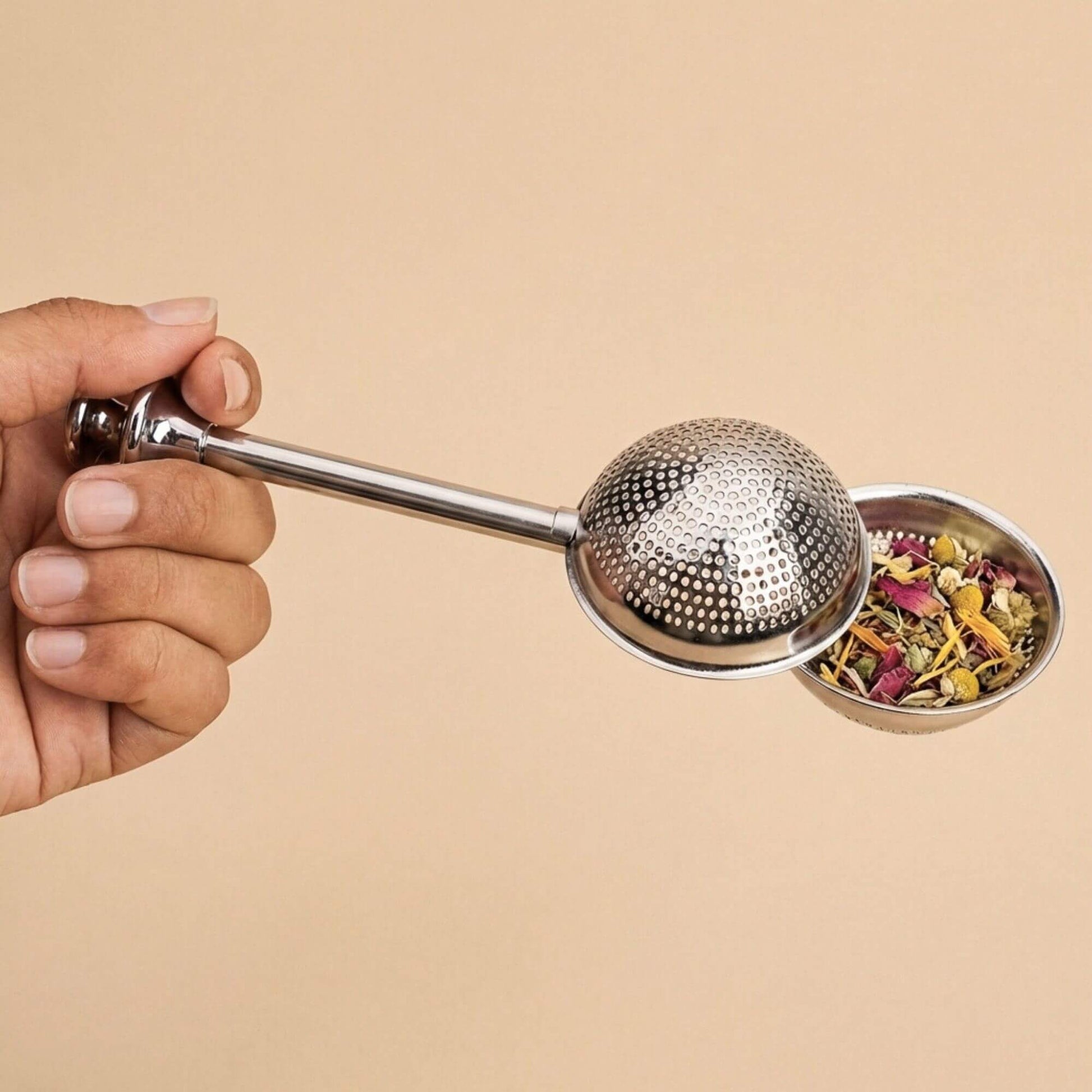 Boule à thé en inox remplie de plantes en vrac, tenue en main, accessoire pour infusion de tisanes et thés sur fond beige.