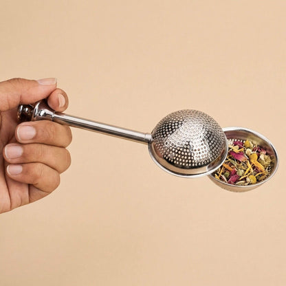 Boule à thé en inox remplie de plantes en vrac, tenue en main, accessoire pour infusion de tisanes et thés sur fond beige.