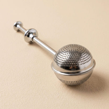 Boule à thé en acier inoxydable fermée, filtre à infusion réutilisable présenté sur fond beige clair.
