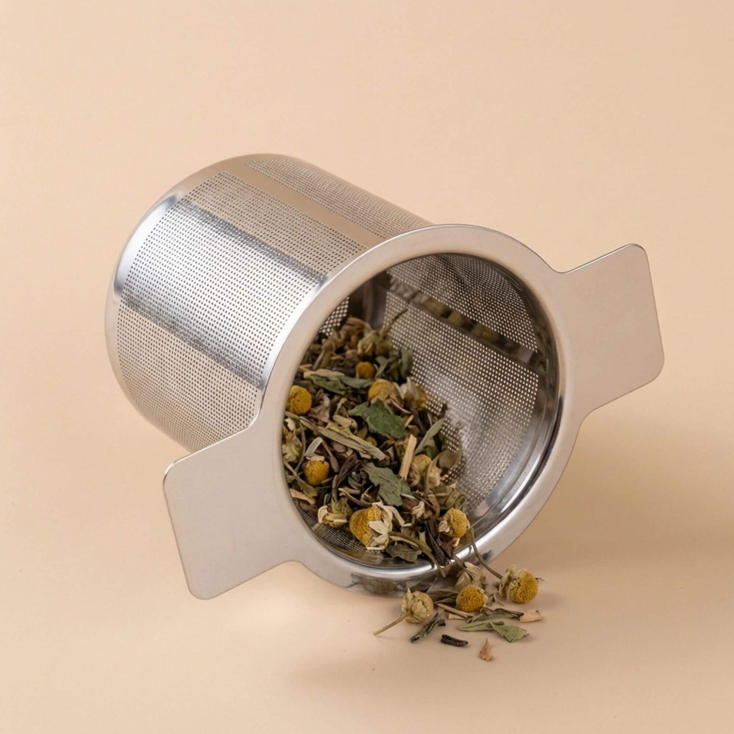 Filtre à thé en acier inoxydable contenant des plantes en vrac pour infusion, présenté incliné sur fond beige.