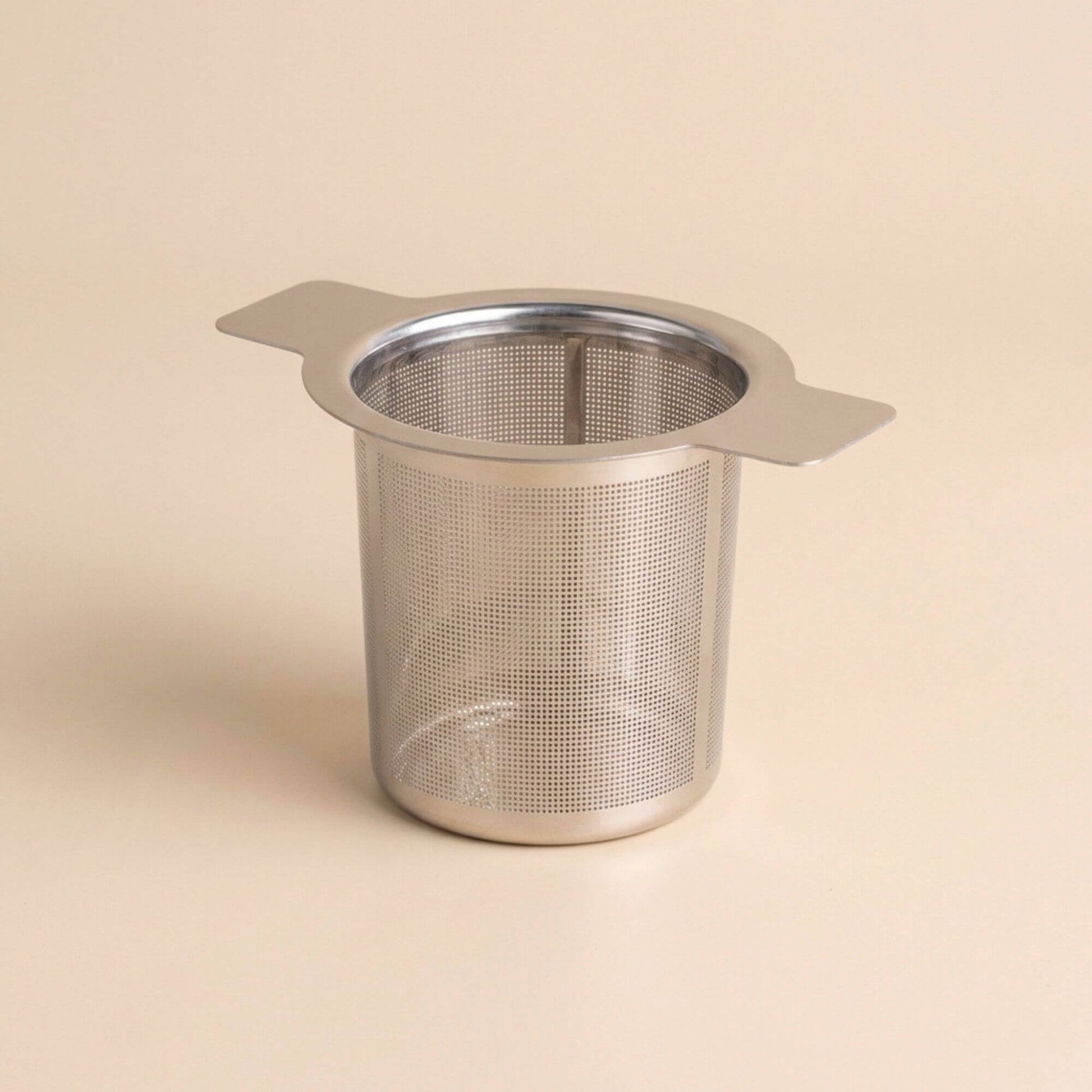 Filtre à thé en inox réutilisable vide, accessoire pour infusion de tisanes et thés, présenté sur fond beige minimaliste.
