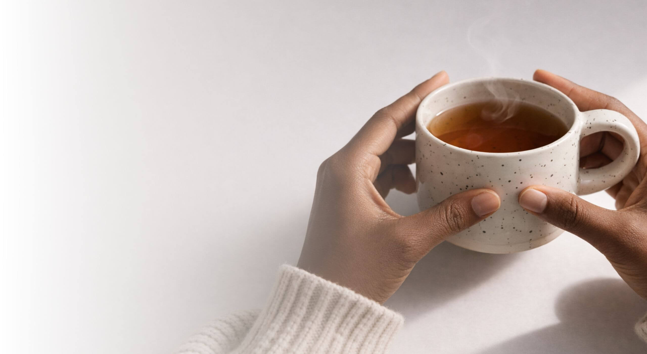 Mains tenant une tasse en céramique beige mouchetée contenant une infusion chaude, avec une vapeur légère et naturelle, dans une ambiance douce et minimaliste évoquant un moment de calme et de bien-être.